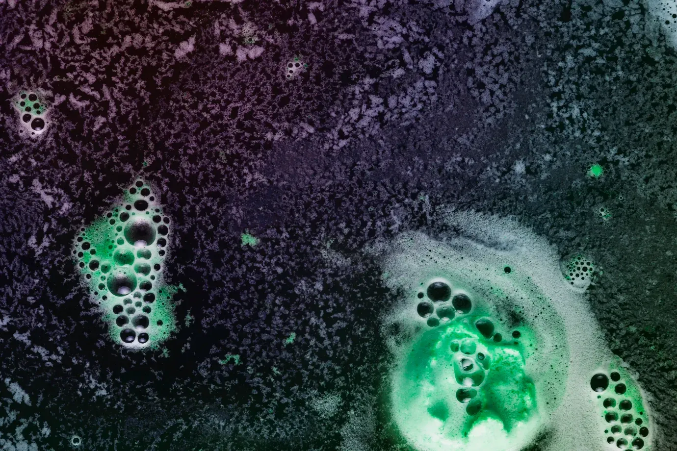 Nahaufnahme von grünlich schimmerndem Schaum auf dunkler, strukturierter Oberfläche. Die Schaumblasen leuchten smaragdgrün und heben sich kontrastreich vom violett-schwarzen Hintergrund ab. Die Szene wirkt chemisch oder experimentell.