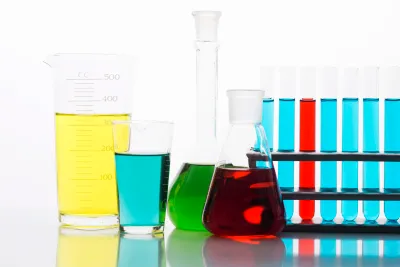Laborgefäße aus Glas mit farbigen Flüssigkeiten: Messbecher, Bechergläser, Erlenmeyerkolben und Reagenzgläser mit gelben, grünen, blauen und roten Lösungen vor weißem Hintergrund.