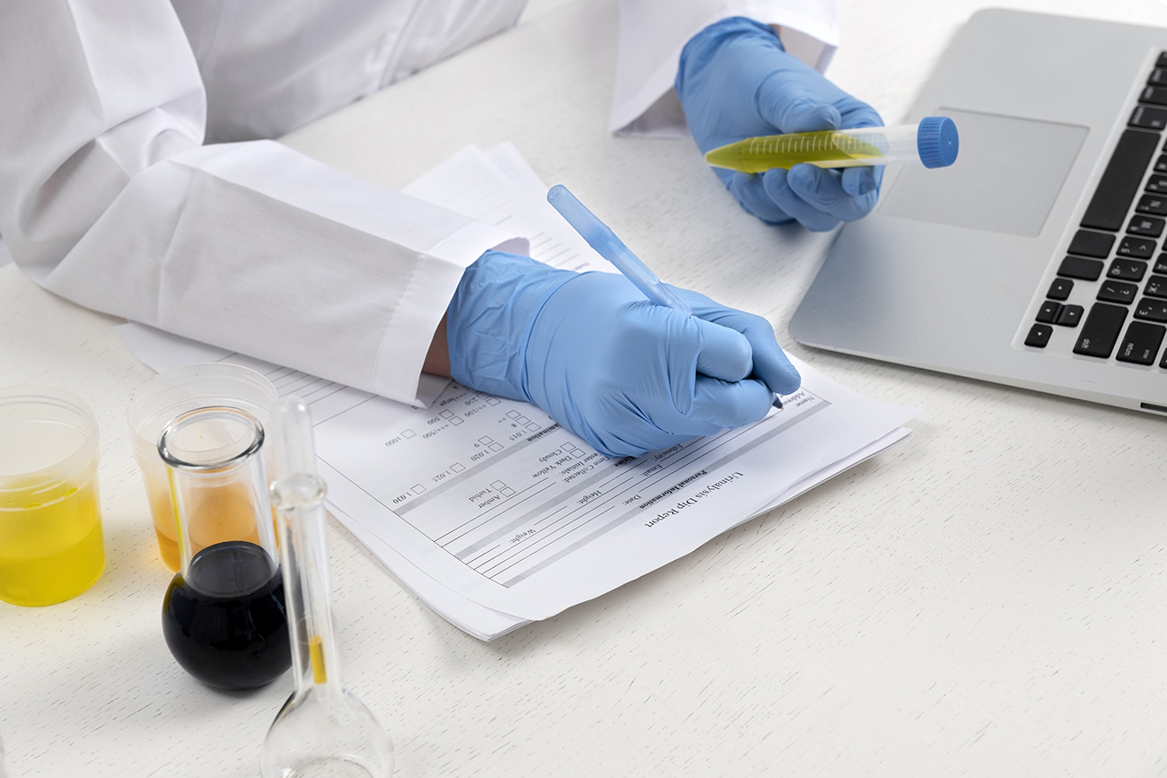 Eine Person in einem weißen Laborkittel und blauen Schutzhandschuhen schreibt auf ein Formular und hält gleichzeitig ein Reagenzglas mit gelber Flüssigkeit. Auf dem Tisch stehen weitere Laborgefäße mit verschiedenfarbigen Flüssigkeiten sowie ein Laptop.