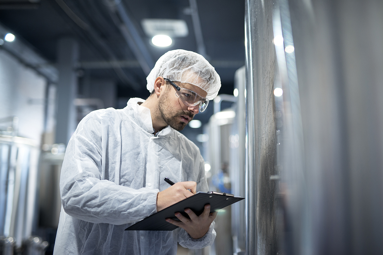 Ein Mitarbeiter in Schutzkleidung, Haarnetz und Schutzbrille inspiziert eine Industrieanlage und macht Notizen auf einem Klemmbrett. Die Umgebung ist eine Produktionsstätte mit Edelstahltanks.