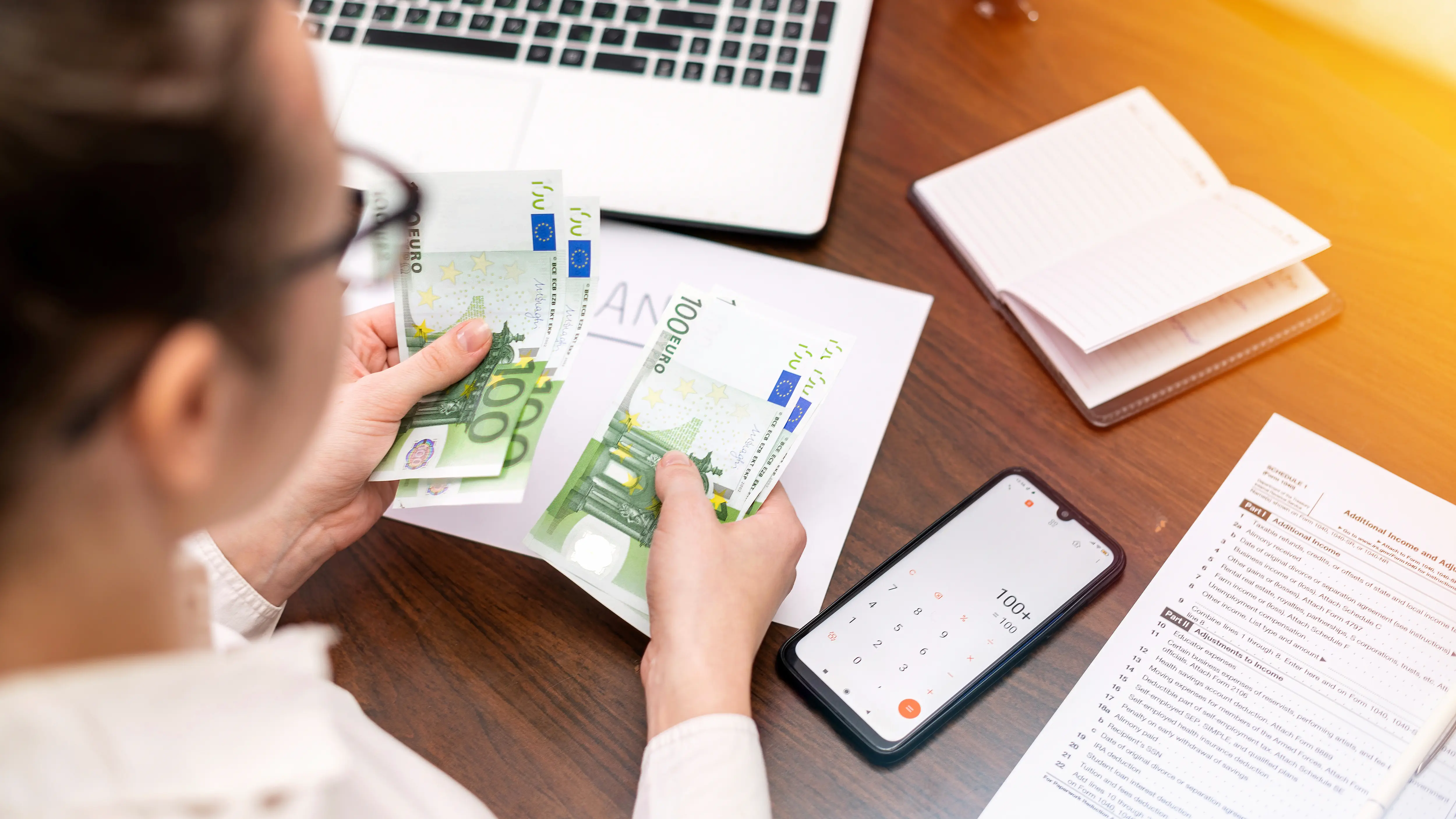 Eine Person zählt mehrere 100-Euro-Scheine an einem Schreibtisch, auf dem ein Laptop, ein Smartphone mit geöffnetem Taschenrechner, ein Notizbuch und Steuerunterlagen liegen.